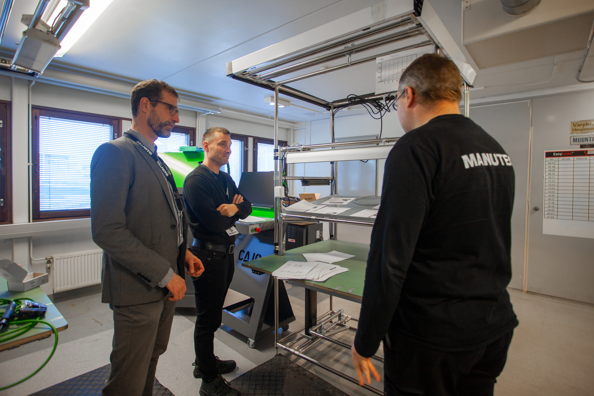 Kolme miestä keskustelevat tuotantosolun vieressä Manutecin tiloissa. Vasemmalla pukupaidassa Jari Vuorinen ja keskellä kädet ristissä Jukka Porkka kuuntelevat, kun oikealla Manutec-paidassa oleva Olli Sulima esittelee tuotantolinjaa.