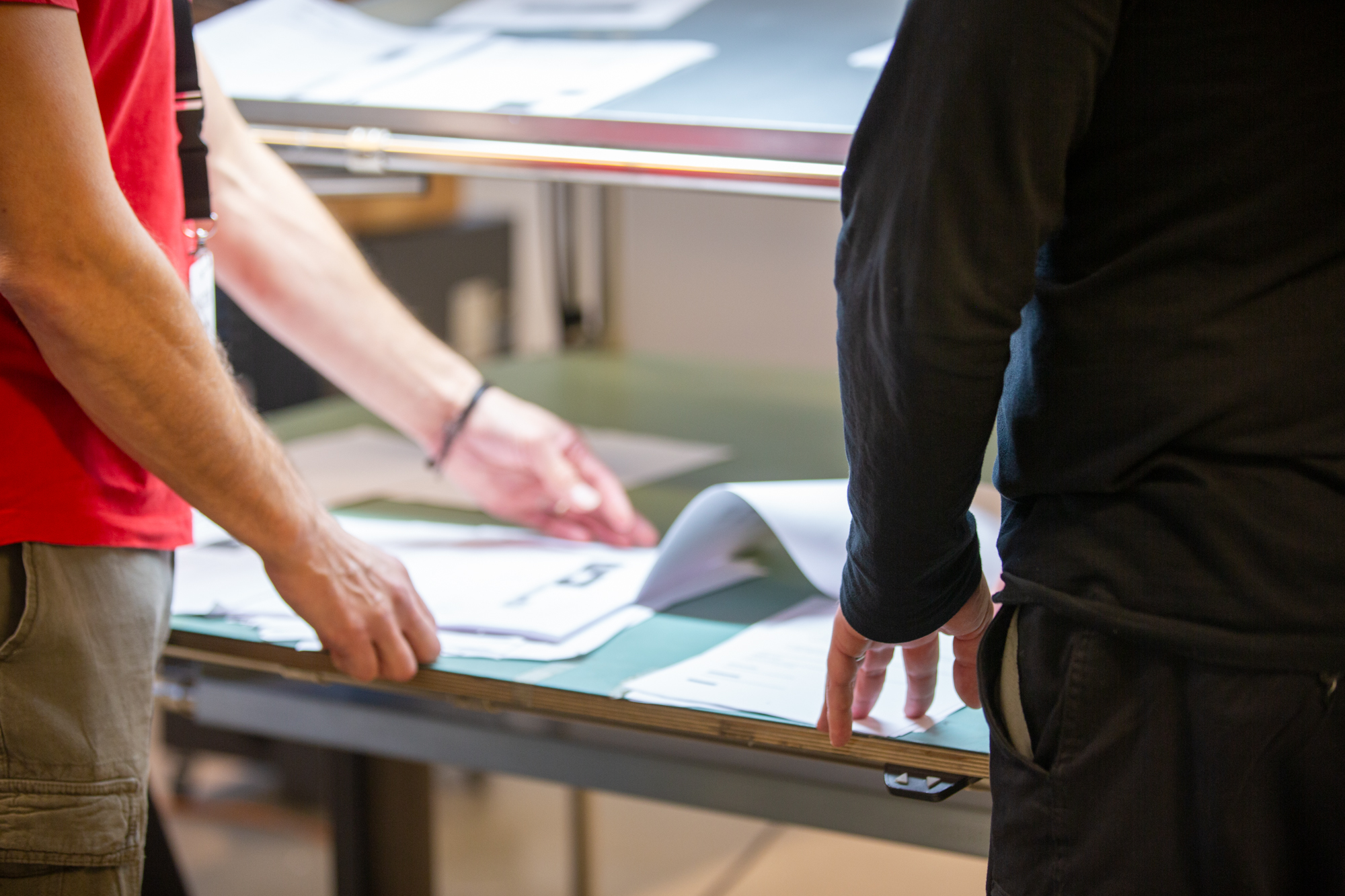 Kaksi henkilöä tarkastelee tuotantosolun piirustuksia pöydän päällä. Vasemmalla punaisessa paidassa ja oikealla mustassa Manutec-paidassa olevat työntekijät osoittavat käsillään suunnitelmia.