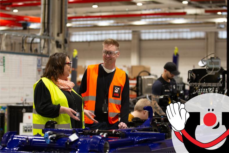 Teollisuushallissa kaksi henkilöä keltaisessa ja oranssissa huomioliivissä keskustelevat sinisen vaihtolavakomponentin äärellä. Kuvassa vasemmalla on Manutecin toimitusjohtaja Kirsi Junnilainen ja oikealla Hiabin Jussi Peltoniemi. Oikeassa alakulmassa näkyy Manutecin Manu-maskottihahmo.